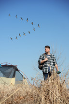 a young man walking outdoors near a tent and geese flying in a "V"