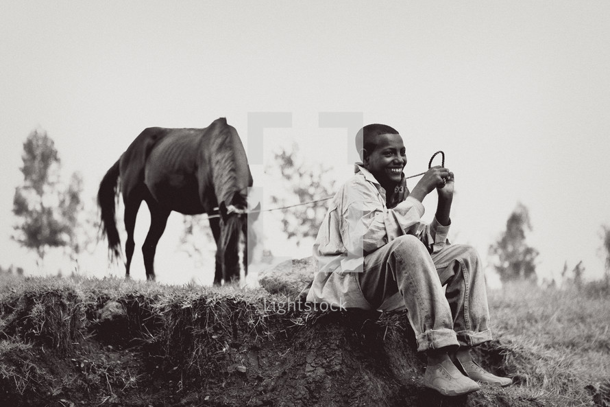 Smiling boy sitting on dirt mound in field with trees holding rope tied to horse.