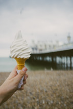 Hand holding an ice cream cone near a pier on a beach.
