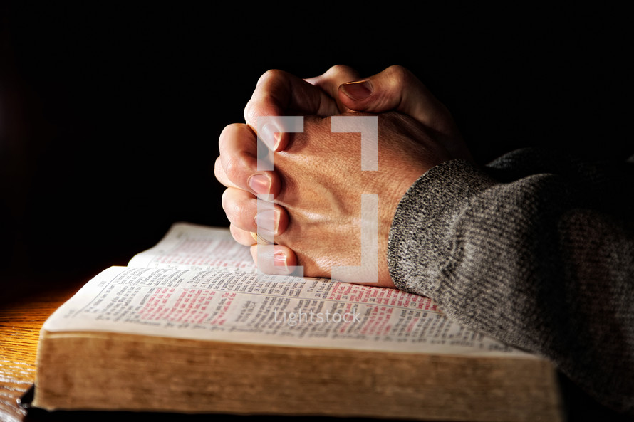A man's hands clasped in deep prayer over a Holy Bible.