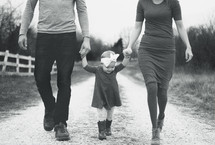 a mother and father walking holding hands with their daughter daughter down a gravel road 