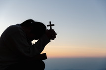 Silhouette of woman with cross praying during sunset