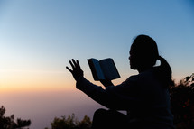 Silhouette of woman with Bible praying during sunset