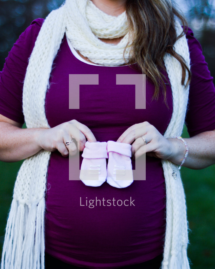 a pregnant woman in a knitted scarf holding baby booties 