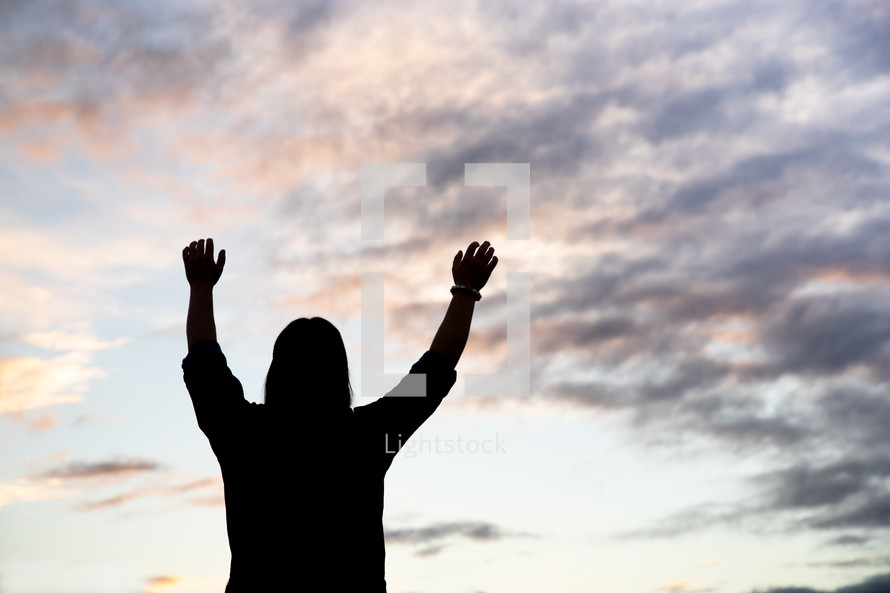 silhouette of a young woman standing outdoors at sunset with hands raised 