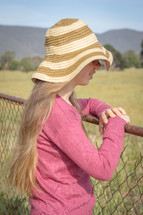 a girl in a straw hat looking over a fence 