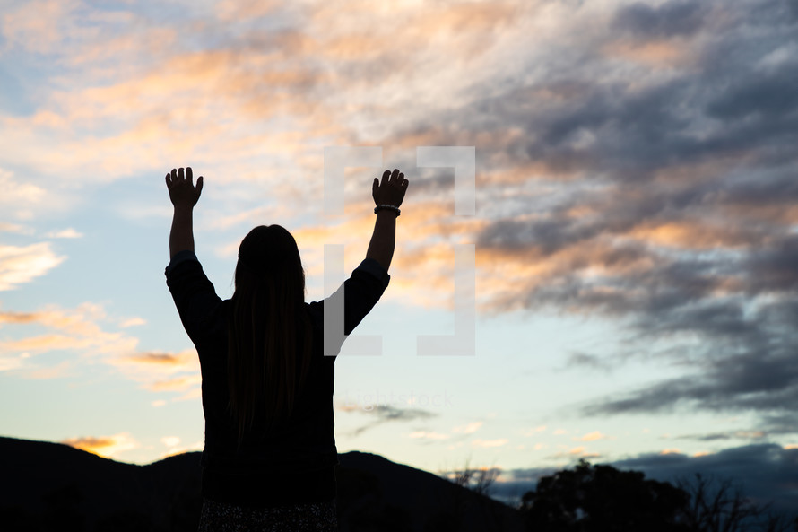 silhouette of a young woman standing outdoors at sunset with hands raised 