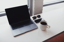 laptop computer, cellphone, coffee cup, and sunglasses on a counter 