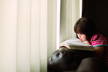 a girl reading a Bible on the arm of a couch 