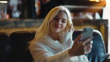 Blonde woman looking at her cell phone and taking a selfie in a modern cafe.