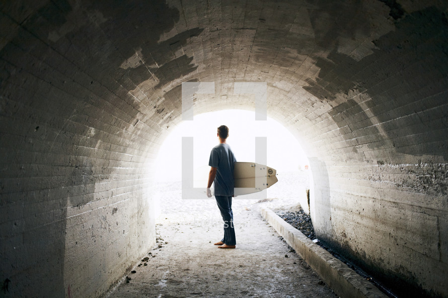 man holding a surf board walking under a tunnel