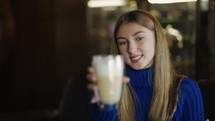 Smiling woman holds big transparent cup of cappuccino in hands then drinks.