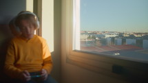 12-year-old teenager sits by the window overlooking the city, wearing white headphones, and sings along to his phone during sunrise. Focus pulling from the window to the boy at the beginning of the clip