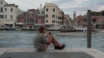 A teenager in a striped shirt is captivated by his phone, while seated on the edge of a Venice canal with boats and historic buildings in the background