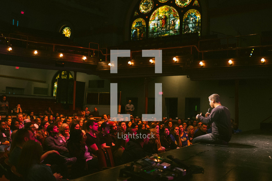 A man sitting on a stage and talking to a congregation in a church sanctuary.