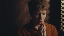 Young faithful female penitent holding hands together and saying a prayer with eyes closed in confessional stall
