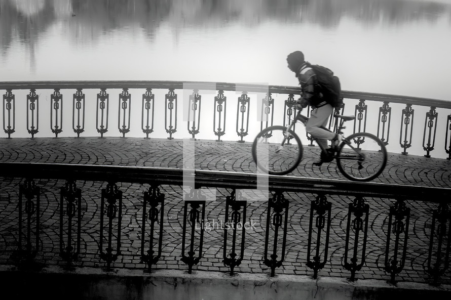Unidentifiable hooded cyclist crossing a paved old bridge. Black and white image
