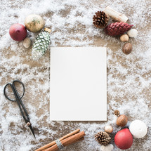 white paper in snow with border of scissors, cinnamon sticks, and pine cones 
