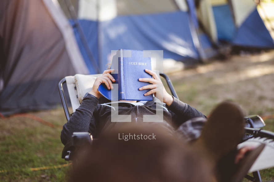 a man camping sitting in a lounge chair reading a Bible 