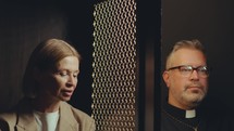 Catholic priest and senior woman sitting on both sides of lattice screen and having open conversation during confession in church
