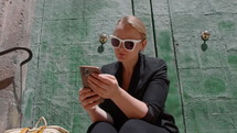 Medium shot of young woman wearing sunglasses with a white frame, sitting outside near a large green door, with a mobile phone in her hand