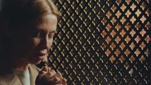 Mature female parishioner praying to God with eyes closed, priest sitting behind lattice screen in confessional booth
