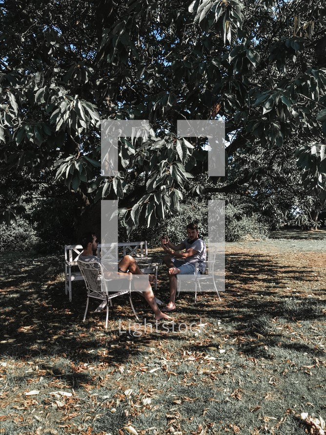 men sitting in chairs in the shade under a tree 