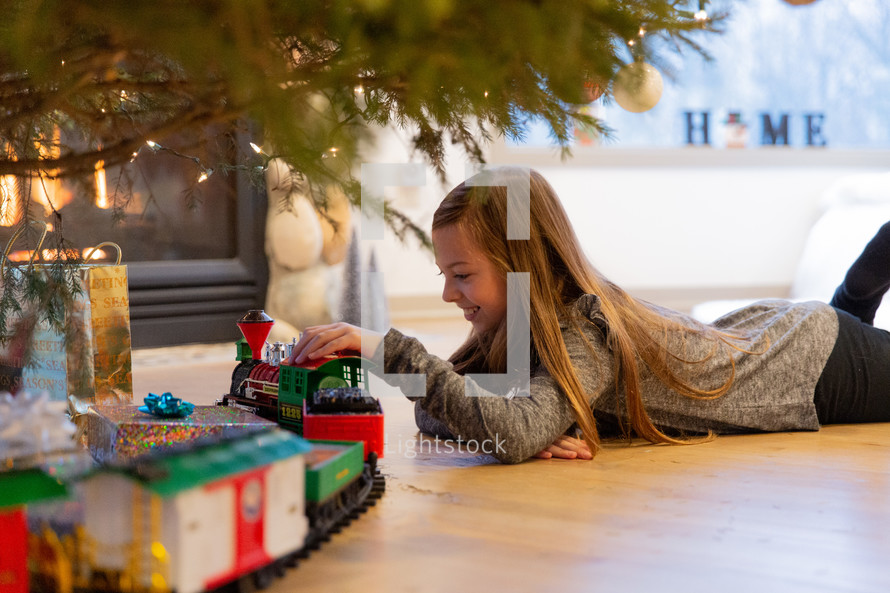little girl playing with a toy train under a Christmas tree