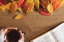 Open bible and hands holding a cup of coffee with autumn leaves on a wood table