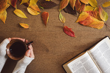Open bible and hands holding a cup of coffee with autumn leaves on a wood table