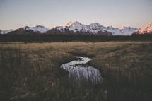 stream running through a field and snow capped mountain peaks at sunset 