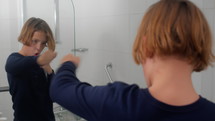 A teenage boy wearing a blue t-shirt is shown boxing in front of a bathroom mirror, practicing his moves and punches