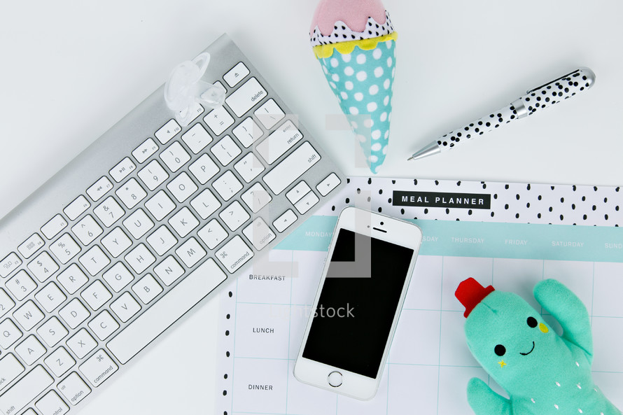 computer keyboard, ice cream cone and cactus plush toy, cellphone, planner, and pen on a desk 
