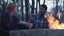 Young man and woman sit beside a fire and make s’mores 
