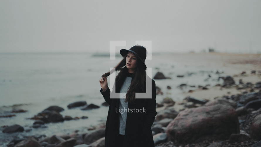 a young woman in a trench coat and hat standing on a rocky shore 