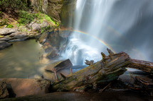 Scene of silky waterfall with rainbow surrounded by mossy rocks and fallen tree logs