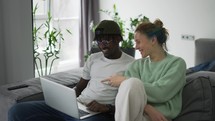 Happy biracial couple sitting on sofa with laptop, congratulating each other.