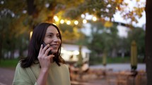 Portrait of a brunette in a green sweater, who speaks by phone against the backdrop of the bright lights of a cafe in the park. Evening walk, talking on the phone.