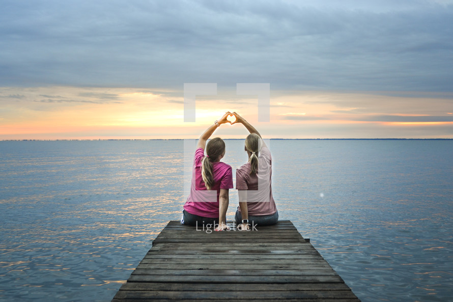 sisters sitting at the end of a dock making a heart with their hands 