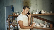 A happy young potter in glasses is talking on a smartphone while standing near his clay works. Traditional business concept.