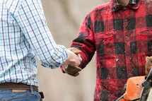 White collar man in formal attire shaking hands with blue collar working outdoorsman holding chainsaw outside