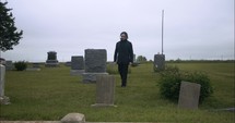 Young, sad man in black suit in cemetery at graveyard tombstone grieving in cinematic slow motion.