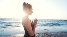 Young girl in black bodysuit practicing yoga with namaste mudra near sea or ocean. Healthy lifestyle concept.
