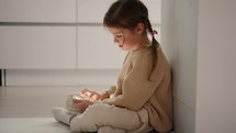 Close-up of a little brown-haired girl in a cream sweater sitting on the floor near the kitchen table and doing something on the phone during her time alone in the evening