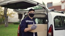 Handsome courier takes cardboard box package out of delivery van walks through private sector. Courier in mask, cap and gloves on the way to deliver postal parcel to a client.