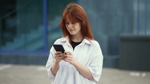a red-haired girl in a shirt is typing text on the phone against the background of a city building.