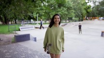 A happy brunette girl in a green sweater walks with a skateboard in the park and smiles. Walk in the park, portrait. Hobby, leisure.
