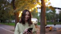 A brunette girl in a green sweater listens to music on headphones against the backdrop of an evening park. Bright lights near the cafe in the park. Evening walk, leisure.