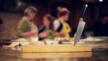 A knife in a cutting board in front of a cooking class.