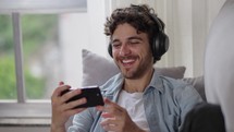 Cheerful young caucasia man watch funny video and laughing wearing headphone. Funny male layinug on the couch at home having fun listening podcast 
